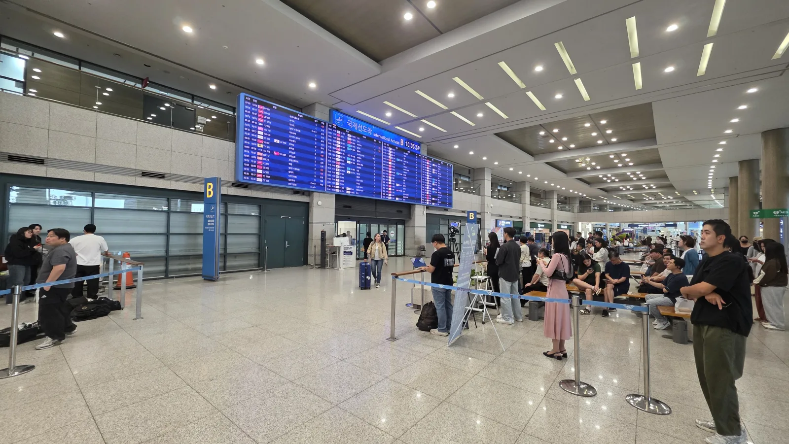 인천공항 국제선 도착 B게이트 전광판 10시 5분, 차단봉 뒤에서 기다리는 사람들과 카메라 삼각대