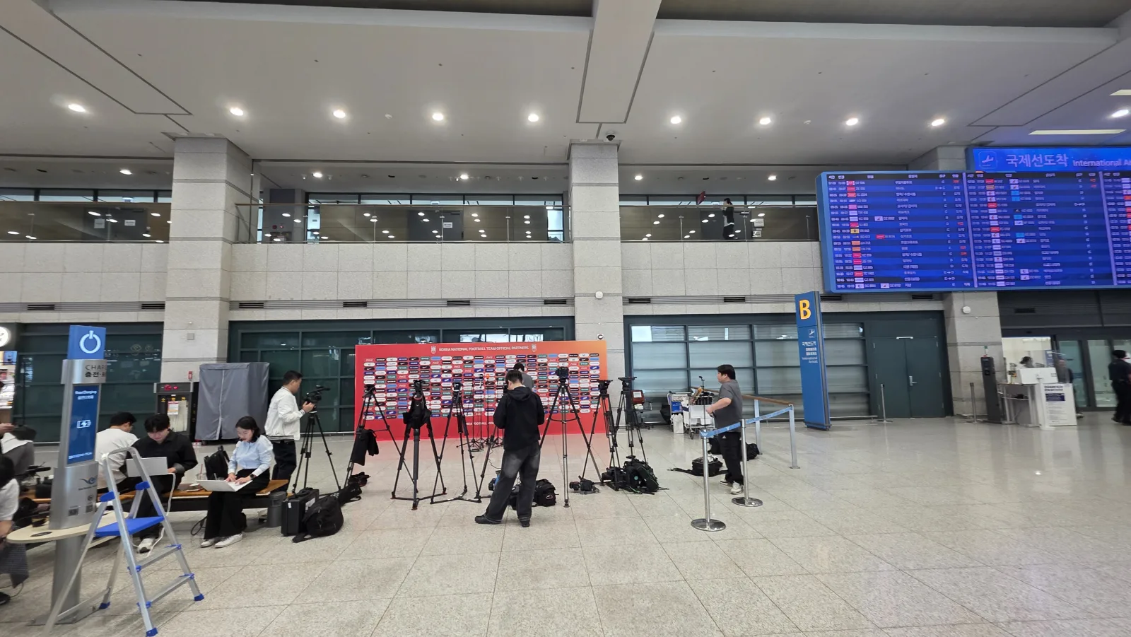 인천공항 도착홀에 세워진 KFA 오피셜 파트너 포토월과 방송 카메라 삼각대, 오른쪽에 국제선 도착 전광판